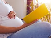 Eine schwangere Person hält eine Broschüre in der Hand, während sie eine Hand auf ihren Bauch gelegt hat